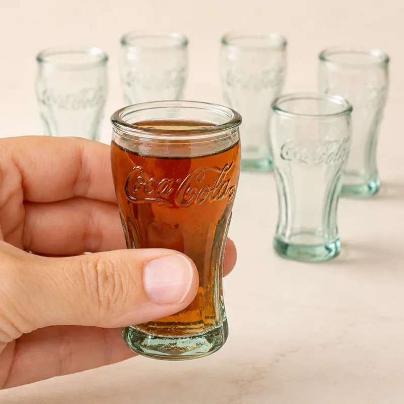 6 Schnapsgläser im Coca Cola Retro Design, eins ist gefüllt und wird von Hand gehalten