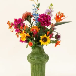 Eine grün strukturierte Vase zeigt den künstlichen Seidenblumenstrauß Colorful Spirit mit Sonnenblumen, rosa Pfingstrosen, orangefarbenen und violetten Blumen und üppigem Grün vor einem schlichten hellen Hintergrund.