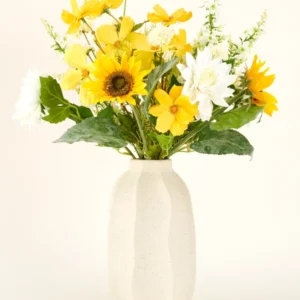 Der künstliche Blumenstrauß Sunny Whisper aus Seide mit Sonnenblumen, weißen Dahlien und grünen Blättern in einer beigen Keramikvase sticht wunderschön vor einem hellen Hintergrund hervor.