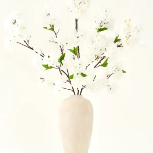 Der künstliche Seidenblumenstrauß Kirschblütencreme 119 cm besteht aus eleganten braunen Zweigen mit zartweißen Kunstblüten und grünen Blättern, die in einer schlichten beigen Vase vor einem schlichten hellen Hintergrund präsentiert werden.