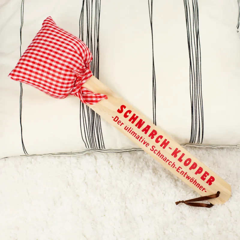 Schnarch-Klopper mit Holzgriff und rot-weiß kariertem Stoffkissen, Aufdruck „Der ultimative Schnarch-Entwöhner“, liegend auf hellem Untergrund
