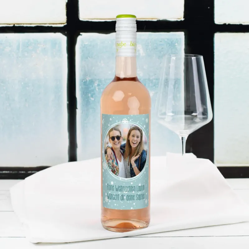 Personalisierte Roséweinflasche mit Schneeflocken-Etikett, eigenem Foto und Weihnachtsgruß – Szene vor Fenster
