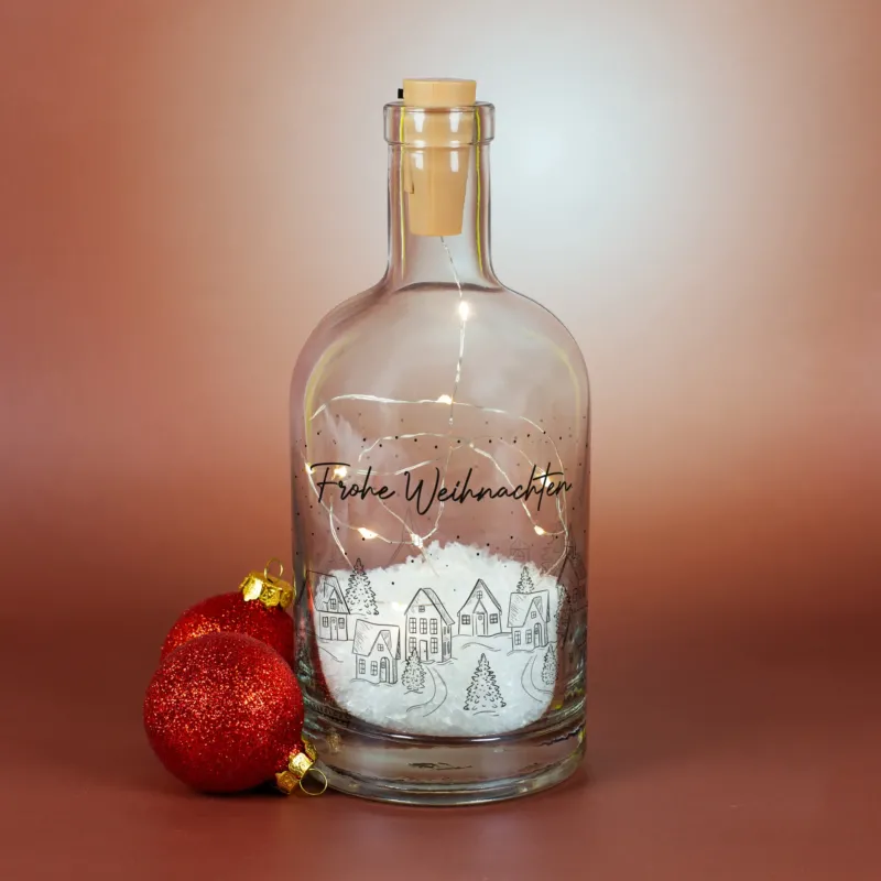Dekorative LED-Karaffe mit Winterdorf-Motiv, Kunstschnee und warmweißer Beleuchtung, personalisiert mit „Frohe Weihnachten“.
