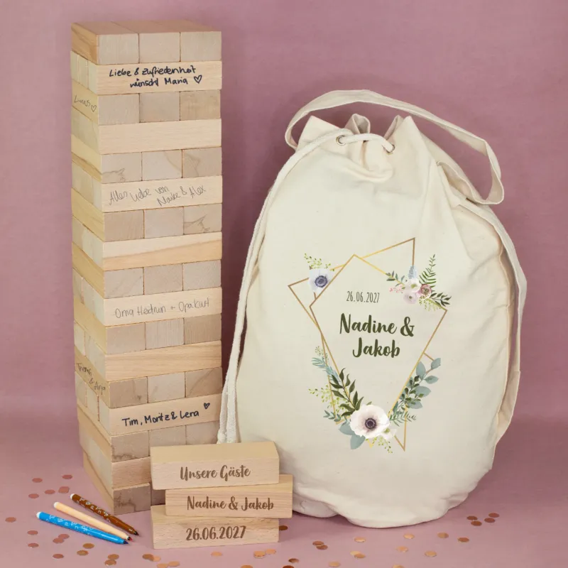 XXL Wackelturm aus Holz als Hochzeitsgästebuch mit beschrifteten Steinen, personalisiertem Seesack mit Blumenmotiv sowie drei gravierten Holzsteinen vor rosafarbenem Hintergrund