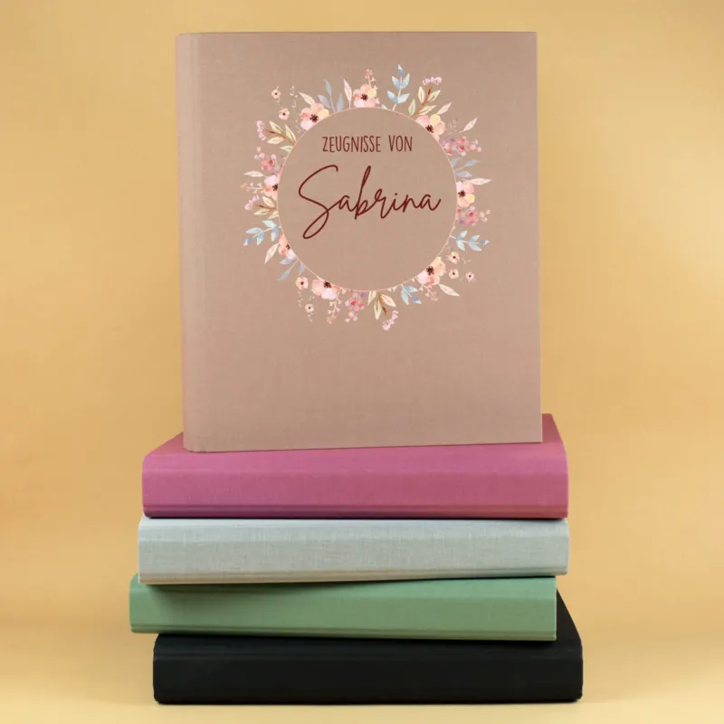 Personalisierte Zeugnismappe aus Leinen in Beige mit Blumenkranz und Name Sabrina auf einem Stapel farbiger Dokumentenmappen.