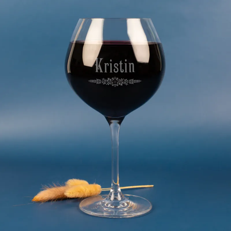 Personalisiertes Leonardo Weinglas mit Gravur des Namens Kristin und floralem Ornament – edles Burgunderglas mit Rotwein, vor blauem Hintergrund mit zarten Trockenhalmen.